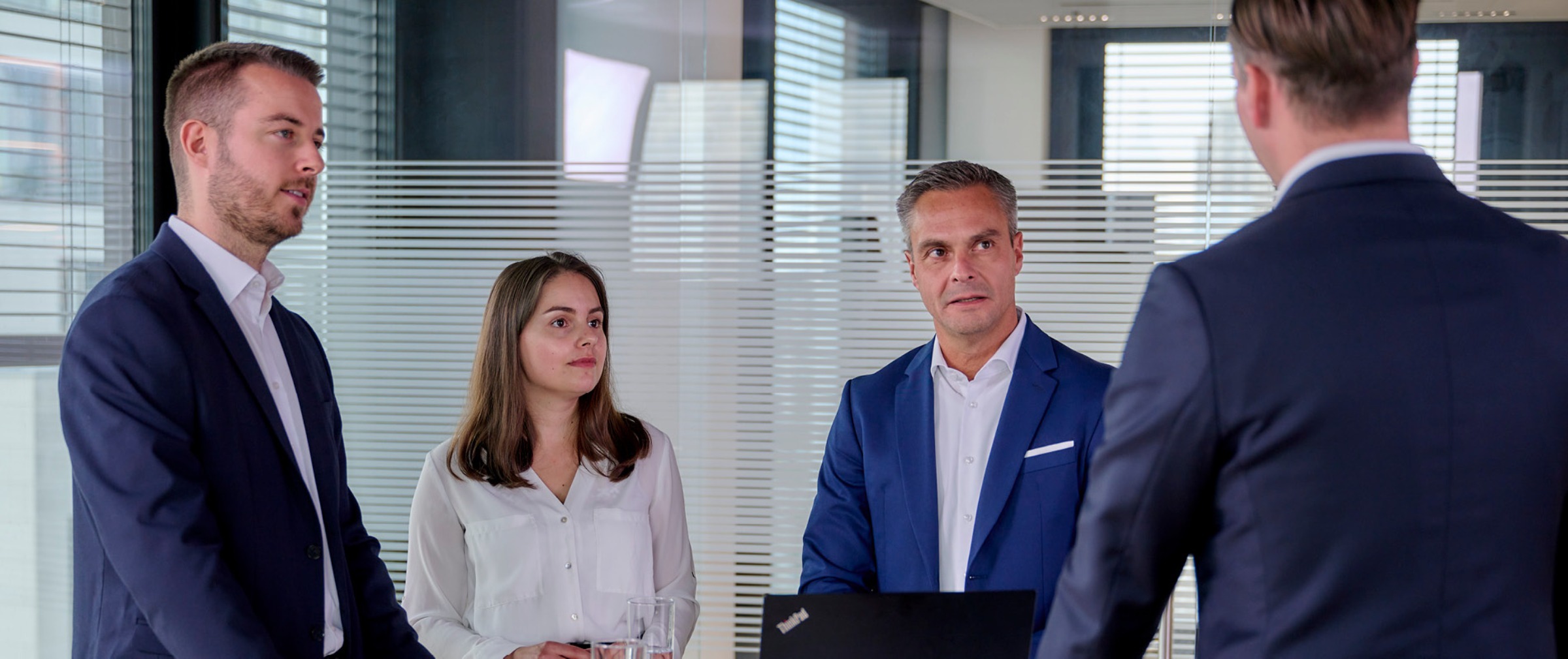 Vier Fondmanager in Businesskleidung führen eine ernsthafte Diskussion in einem modernen Büro mit Glaswänden. Die Stimmung ist fokussiert und nachdenklich.