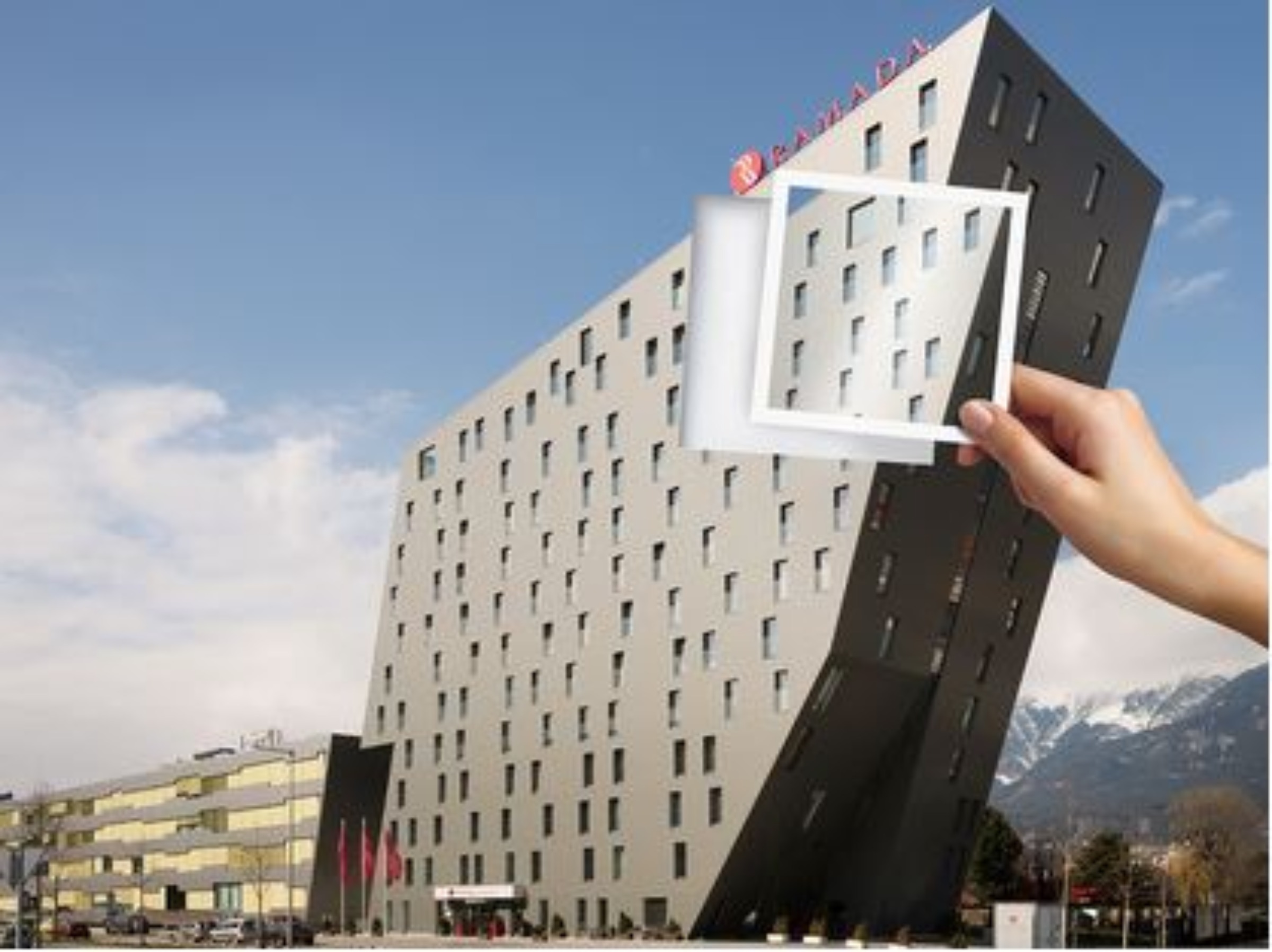 Ein geneigtes modernes Hotel mit kleinen Fenstern vor einem blauen Himmel. Eine Hand hält einen quadratischen Ausschnitt, der verspielt zur Fassade des Gebäudes passt. Im Hintergrund sind schneebedeckte Berge sichtbar.