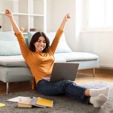 Eine junge Frau sitzt im Wohnzimmer auf dem Boden vor der Couch. Sie lernt gerade und ihr Laptop liegt auf ihren Beinen. 